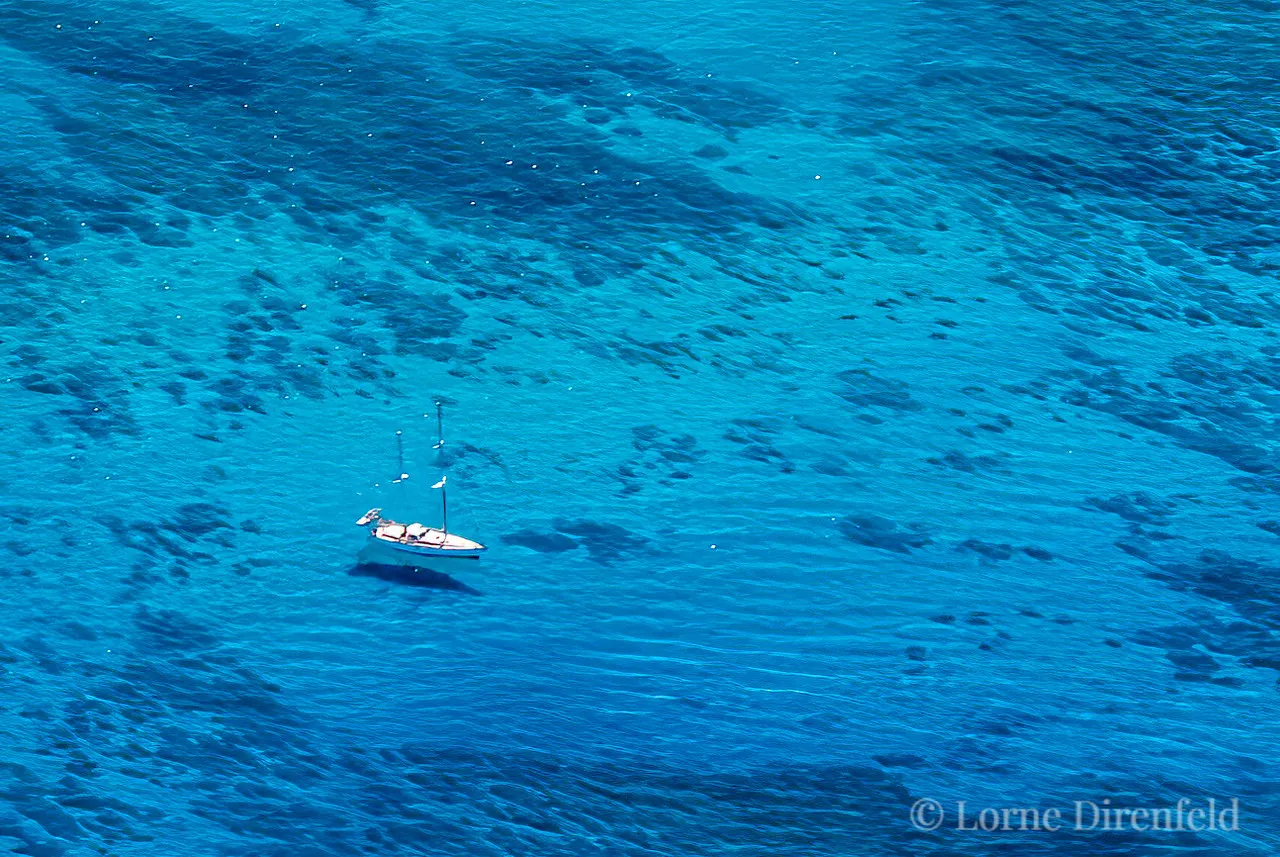 The surfers appear as small dots on the ocean as a sail boat hangs out in the morning in the shimmering blue waters off Waikiki . . .